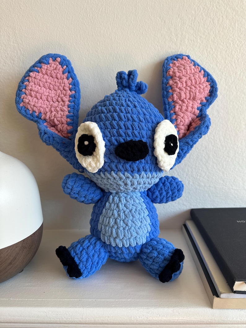 May include: A handmade crochet plush toy featuring a blue alien character with large, upright ears that have pink inner sections. The figure has white eye patches with black pupils, a black nose, and a light blue belly patch with black feet.