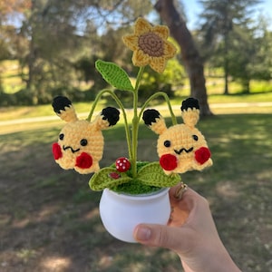 May include: A small white ceramic pot containing a crocheted arrangement with a central yellow sunflower, two yellow Pikachu-like figures with black ear tips and red cheeks, a small red mushroom, and a tiny ladybug, all set on green yarn foliage.