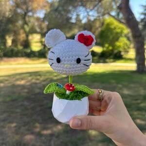 May include: A white crocheted cat head with a red bow, black eyes, and whiskers, attached to a green stem. It sits in a white geometric pot with green crocheted leaves and a small red and white mushroom accent at the base of the stem.