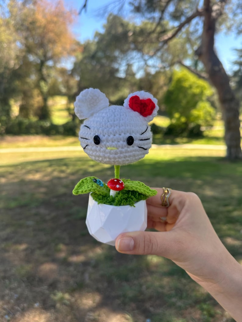 May include: A white crocheted cat head with a red bow, black eyes, and whiskers, attached to a green stem. It sits in a white geometric pot with green crocheted leaves and a small red and white mushroom accent at the base of the stem.