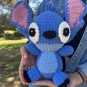 May include: A blue crochet plush toy resembling the character Stitch, featuring large upright ears with pink inner sections, white oval eyes with black pupils, a black nose, and a light blue belly patch, crafted from thick, soft yarn.