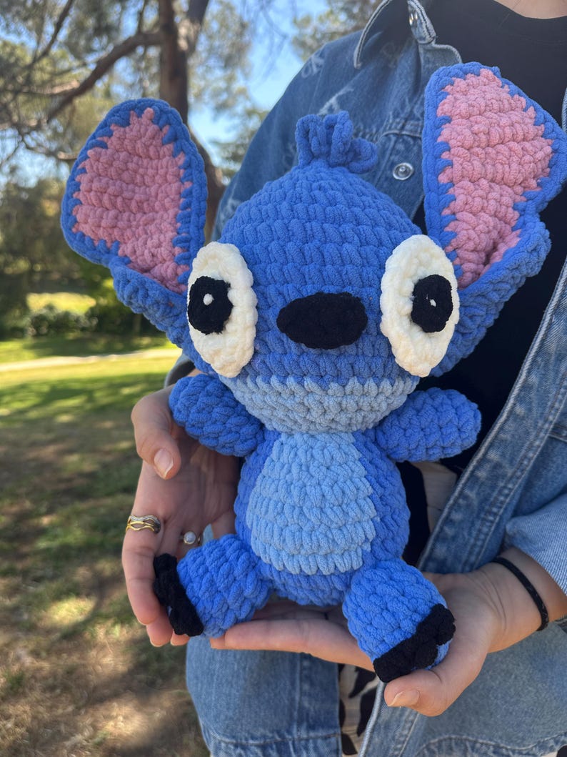 May include: A blue crochet plush toy resembling the character Stitch, featuring large upright ears with pink inner sections, white oval eyes with black pupils, a black nose, and a light blue belly patch, crafted from thick, soft yarn.