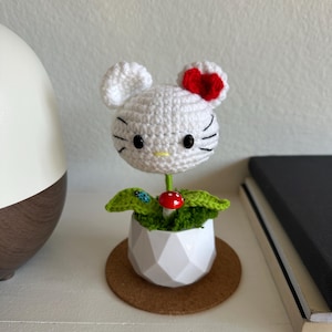 May include: A white crocheted character head with black eyes, whiskers, and a red bow, attached to a green stem. It sits in a white geometric pot with green crochet leaves, a small red mushroom, and a tiny blue ladybug accent on one leaf.