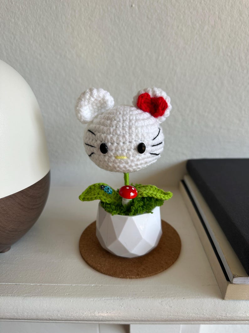 May include: A white crocheted character head with black eyes, whiskers, and a red bow, attached to a green stem. It sits in a white geometric pot with green crochet leaves, a small red mushroom, and a tiny blue ladybug accent on one leaf.