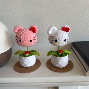 May include: Two crocheted figures shaped like cat heads, one pink and one white, each featuring a red bow, black eyes, and whiskers. They are mounted on green stems inside white geometric pots, decorated with small red mushrooms and green leaves.