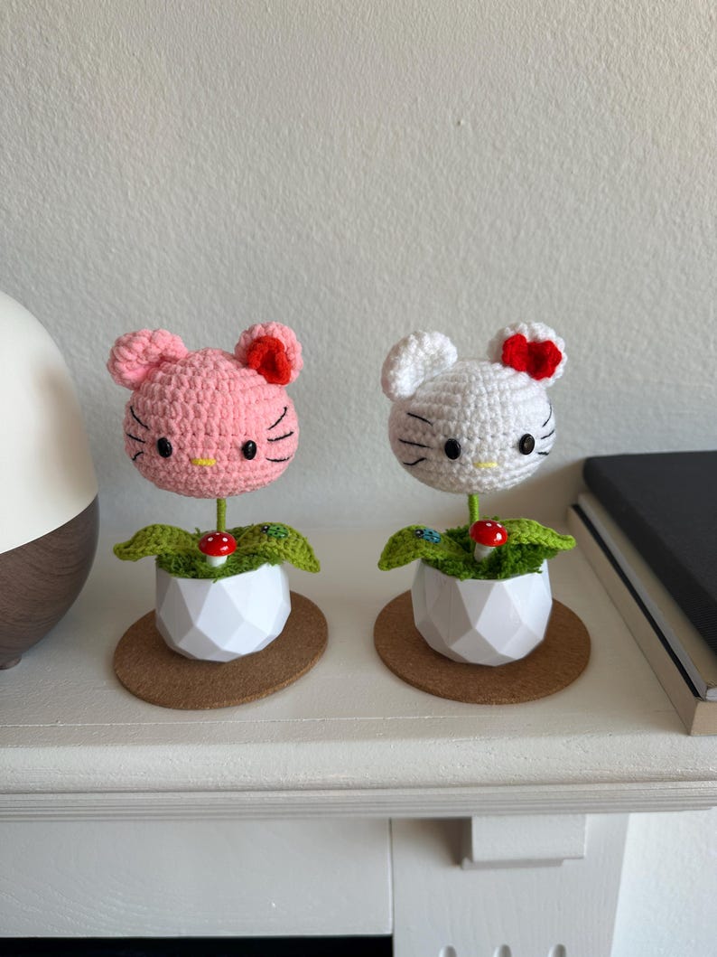 May include: Two crocheted figures shaped like cat heads, one pink and one white, each featuring a red bow, black eyes, and whiskers. They are mounted on green stems inside white geometric pots, decorated with small red mushrooms and green leaves.