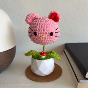 May include: A crocheted pink cat-shaped head with black eyes and whiskers, a yellow nose, and a red ear accent, mounted on a green stem. The base is a white geometric pot containing green crocheted leaves, a small red mushroom, and a ladybug.