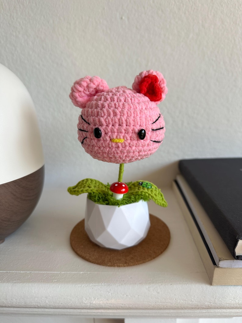 May include: A crocheted pink cat-shaped head with black eyes and whiskers, a yellow nose, and a red ear accent, mounted on a green stem. The base is a white geometric pot containing green crocheted leaves, a small red mushroom, and a ladybug.