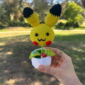 May include: A yellow crocheted character with long ears tipped in black, black eyes, and red cheeks, attached to a green stem. It sits in a small white geometric pot with green moss, a tiny red and white mushroom, and a small blue ladybug charm.