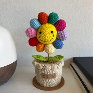 May include: A crocheted flower with a bright yellow smiley face center and multi-colored petals in red, orange, light blue, pink, green, purple, and cream. The flower sits in a beige crocheted pot with a brown tag labeled Hand Made.