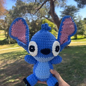 May include: A blue crochet plush toy resembling the character Stitch, featuring large upright ears with pink inner sections, white eye patches with black pupils, a black nose, and a light blue belly patch, crafted from soft, chunky yarn.