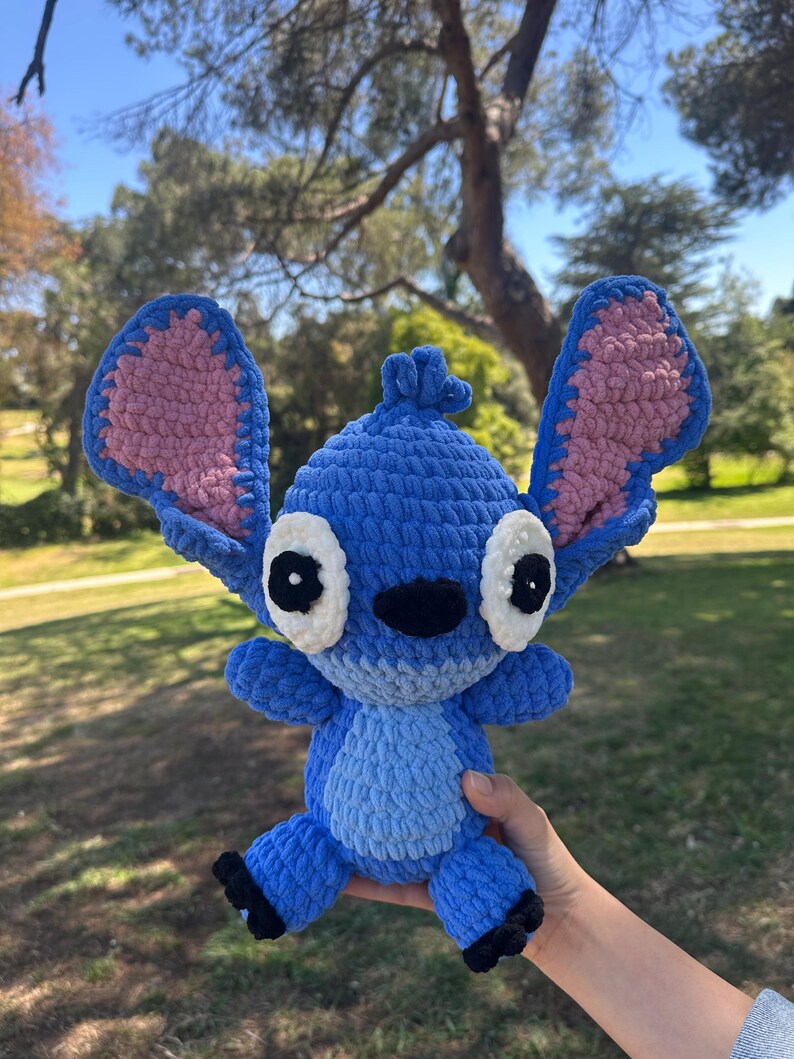 May include: A blue crochet plush toy resembling the character Stitch, featuring large upright ears with pink inner sections, white eye patches with black pupils, a black nose, and a light blue belly patch, crafted from soft, chunky yarn.