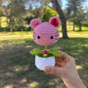 May include: A pink crocheted cat-shaped flower head with black embroidered whiskers and eyes, a yellow nose, and a red inner ear. It sits in a white geometric pot with green crocheted leaves, a small red mushroom, and a tiny green ladybug accent.