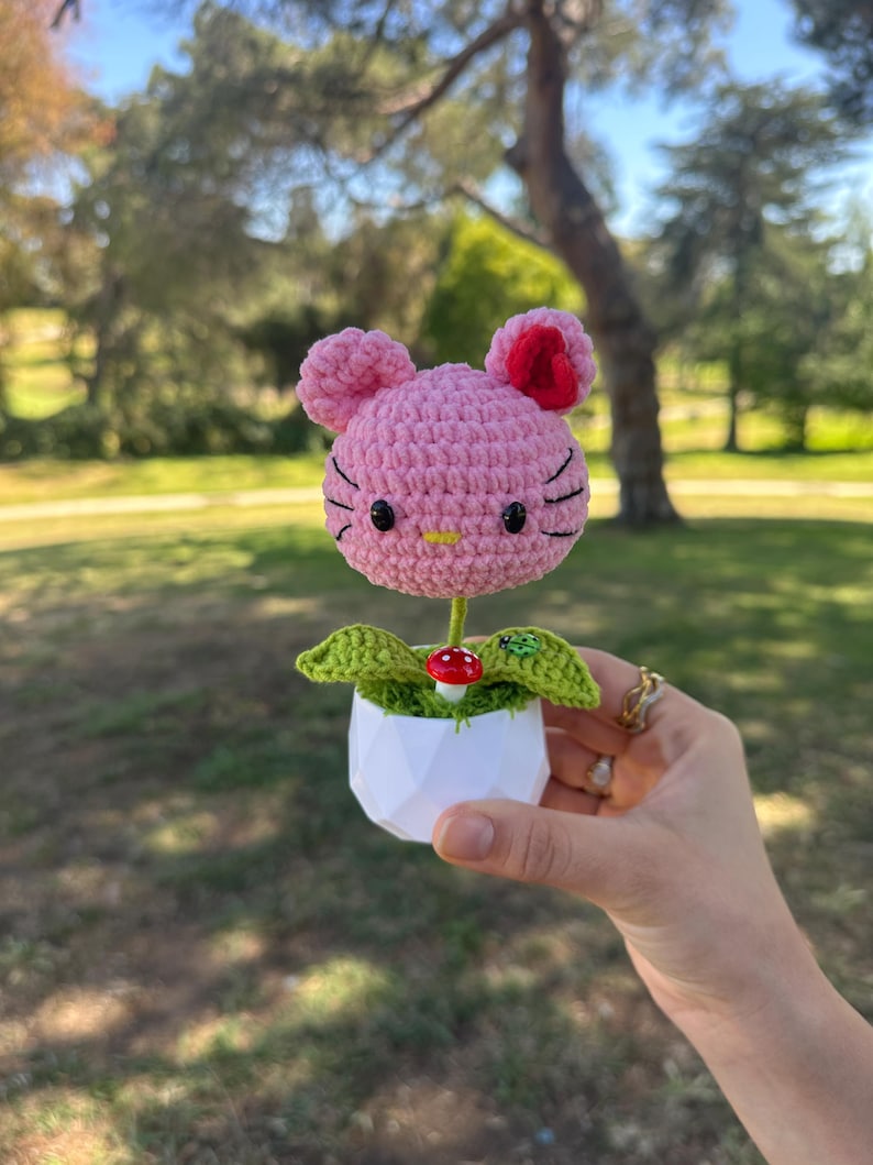 May include: A pink crocheted cat-shaped flower head with black embroidered whiskers and eyes, a yellow nose, and a red inner ear. It sits in a white geometric pot with green crocheted leaves, a small red mushroom, and a tiny green ladybug accent.