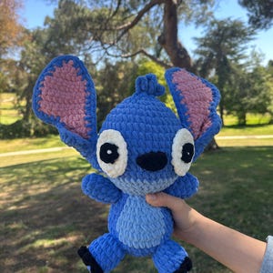 May include: A blue crochet plush toy featuring large upright ears with pink inner sections, white eye patches with black pupils, a black nose, and a light blue belly patch. The character has a textured yarn finish and small black feet.