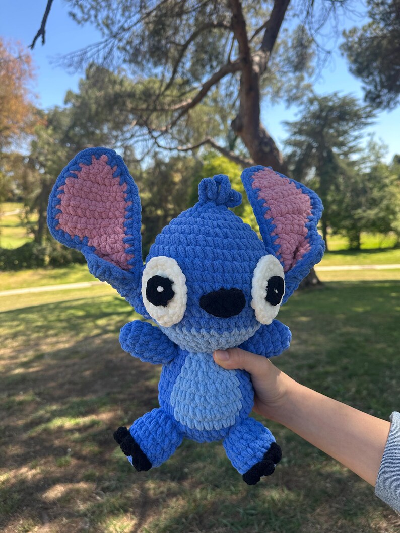 May include: A blue crochet plush toy featuring large upright ears with pink inner sections, white eye patches with black pupils, a black nose, and a light blue belly patch. The character has a textured yarn finish and small black feet.