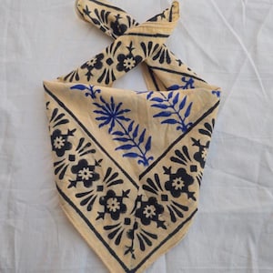 May include: A beige bandana featuring a black floral border and blue and black floral and leaf patterns. The bandana is knotted at the top, displaying the detailed design and color scheme.
