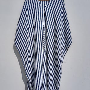 May include: A blue and white striped caftan hangs from a wooden hanger. The caftan has a V-neck and a button-up front. The fabric is a lightweight material with vertical stripes. The garment is a loose, flowing style.