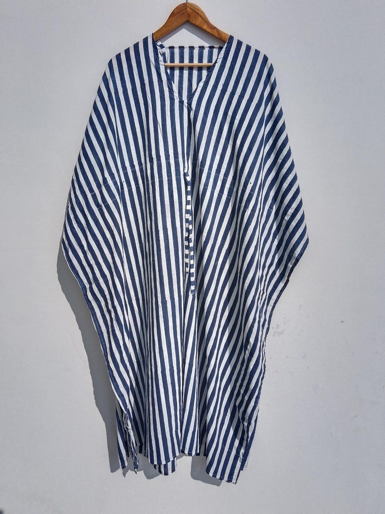 May include: A blue and white striped caftan hangs from a wooden hanger. The caftan has a V-neck and a button-up front. The fabric is a lightweight material with vertical stripes. The garment is a loose, flowing style.