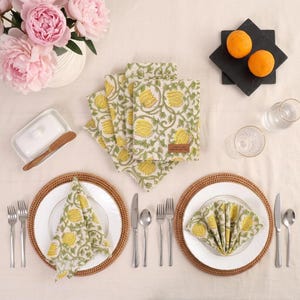 Puede incluir: Un juego de servilletas de algodón con un estampado floral amarillo sobre fondo crema, dispuestas sobre una mesa con platos blancos, manteles individuales de ratán, cubiertos de acero inoxidable, dos naranjas sobre una baldosa negra y un jarrón con peonías rosas.