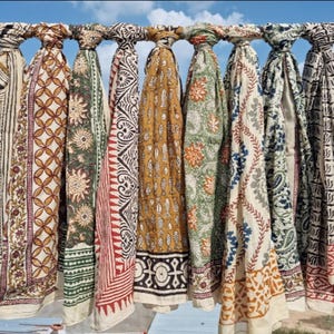 May include: A collection of patterned scarves in various colors, including beige, brown, green, and red, hanging on a metal bar. Each scarf features unique geometric and floral designs, with a mix of light and dark tones.
