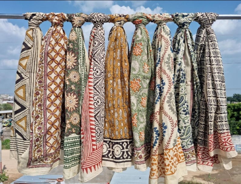May include: A collection of patterned scarves in various colors, including beige, brown, green, and red, hanging on a metal bar. Each scarf features unique geometric and floral designs, with a mix of light and dark tones.