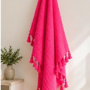 May include: A vibrant hot pink throw blanket featuring a textured cable knit pattern throughout the fabric. The blanket is finished with numerous matching pink tassels attached along the bottom and side edges, hanging in a decorative arrangement.