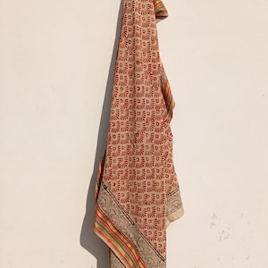 Op de afbeelding: Beige sjaal met een herhalend rood bloemenpatroon en een decoratieve rand. De rand heeft zwarte, gele, rode en groene strepen. De sjaal hangt tegen een witte muur.