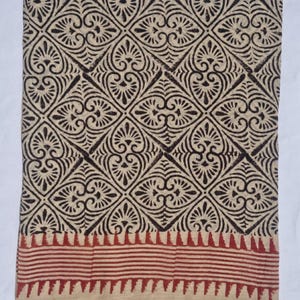 May include: A folded textile with a repeating geometric pattern in dark brown on a beige background. The pattern features diamond shapes with floral-like motifs. A red and beige striped border is at the bottom. The fabric appears to be cotton or a similar natural fiber.