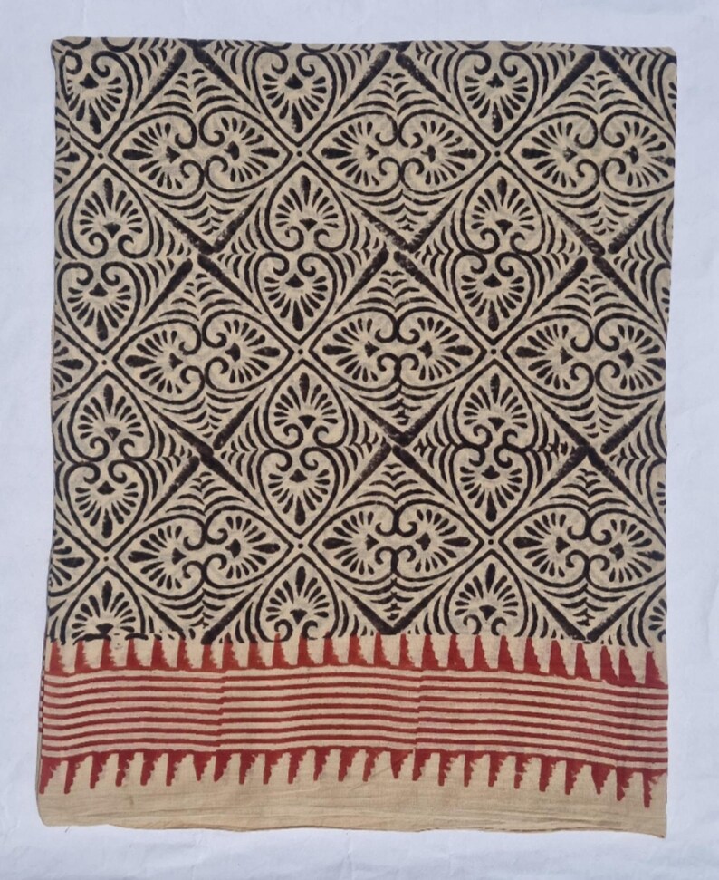 May include: A folded textile with a repeating geometric pattern in dark brown on a beige background. The pattern features diamond shapes with floral-like motifs. A red and beige striped border is at the bottom. The fabric appears to be cotton or a similar natural fiber.