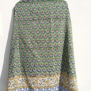 May include: A green textile with a repeating pattern of teardrop-shaped motifs in yellow and blue. The border features a floral design in yellow, white, and blue. The fabric is draped, showcasing the intricate print.