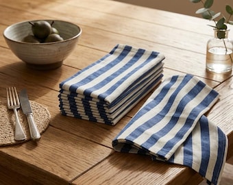 May include: A stack of folded rectangular cloth napkins featuring a pattern of thick navy blue and white vertical stripes. The napkins are arranged on a wooden surface alongside a single unfolded napkin displaying the same blue and white striped design.