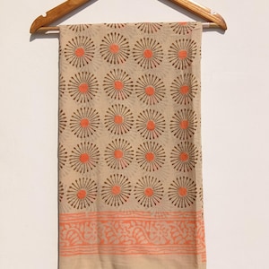 Puede incluir: Tela beige con un patrón repetitivo de diseños de rayos de sol naranjas y marrones. El borde inferior presenta un borde floral naranja. La tela está colgada de una percha de madera, mostrando su textura y patrón.