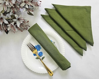 May include: Olive green linen napkins, folded and arranged with a white plate featuring blue floral accents and a gold fork. Decorative white flowers with red accents are in the upper left corner. The napkins are a rich, solid color.