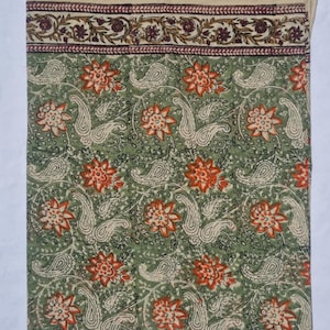 May include: A green textile with a floral and paisley pattern. The design features orange flowers and off-white paisley shapes on a green background. A border with floral and leaf patterns in burgundy and brown accents the top edge.