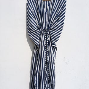 May include: A navy blue and white striped kaftan dress hanging on a wooden hanger. The dress has a V-neck and a drawstring waist. The fabric is lightweight and appears to be cotton or a similar material. The stripes are vertical and evenly spaced.