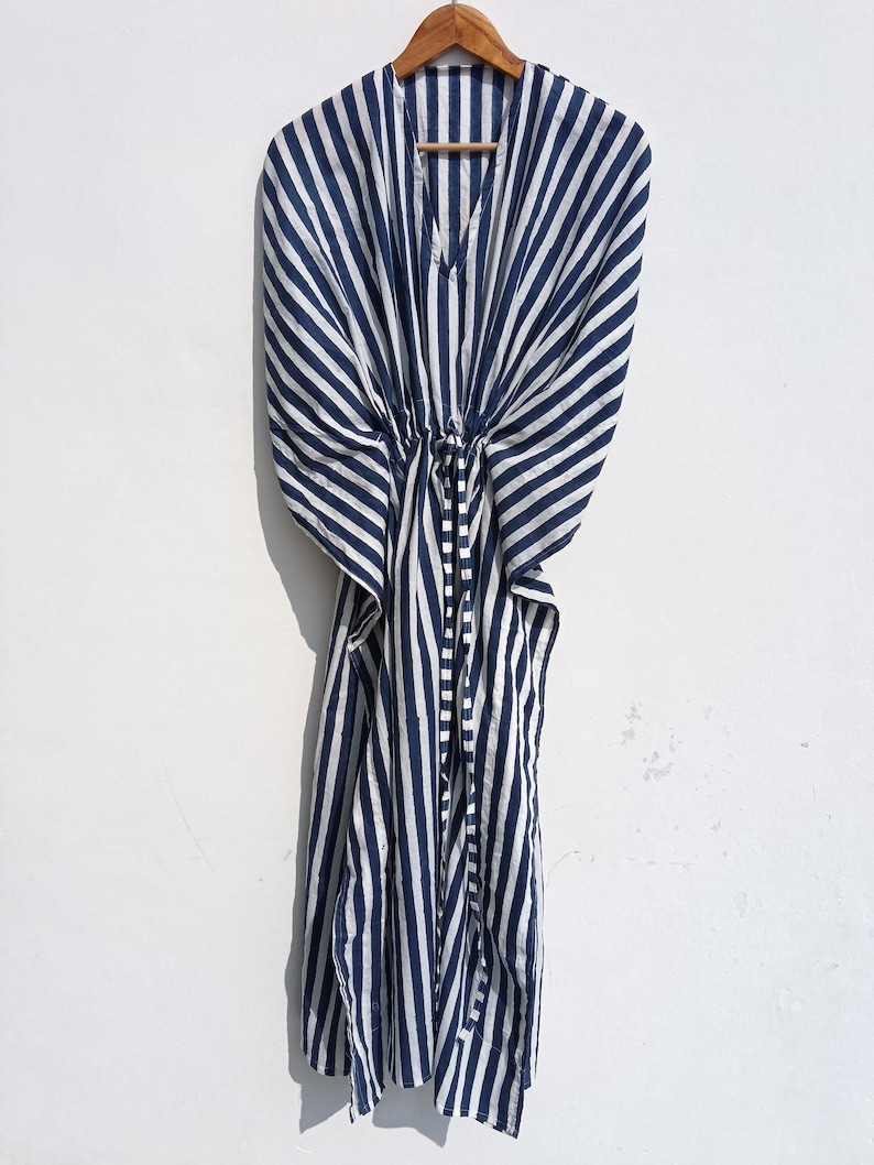 May include: A navy blue and white striped kaftan dress hanging on a wooden hanger. The dress has a V-neck and a drawstring waist. The fabric is lightweight and appears to be cotton or a similar material. The stripes are vertical and evenly spaced.