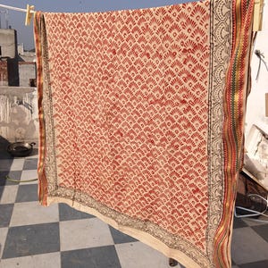 Könnte beinhalten: Ein rechteckiges Textil mit einem sich wiederholenden roten fächerförmigen Muster auf cremefarbenem Grund. Der Stoff ist mit einem schwarzen Blumenmotiv und einem dekorativen Randstreifen mit dünnen goldenen, grünen und roten Streifen versehen.