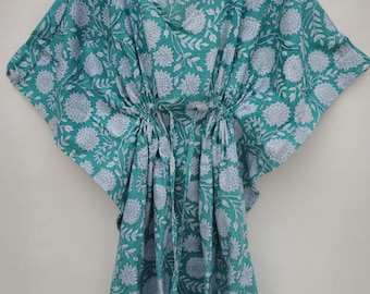 May include: A teal-colored kaftan with a white floral pattern, hanging on a wooden hanger. The kaftan features a V-neck and a drawstring waist. The fabric is lightweight and appears to be cotton or a similar material.
