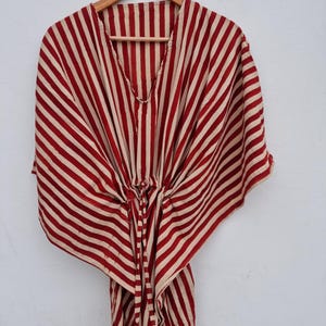 Puede incluir: Un vestido caftán a rayas rojas y beige colgado de una percha de madera. El vestido presenta un escote en V y una cintura con cordón. La tela tiene un patrón de rayas verticales audaz.