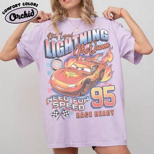 Puede incluir: Una camiseta de color lila claro con un dise&ntilde;o de Lightning McQueen de la pel&iacute;cula Cars. El dise&ntilde;o incluye el texto "Street Legend LIGHTNING McQueen", "Need for Speed" y "Race Ready", junto con el n&uacute;mero "95" y banderas a cuadros. La camiseta est&aacute; etiquetada como "Comfort Colors Orchid".
