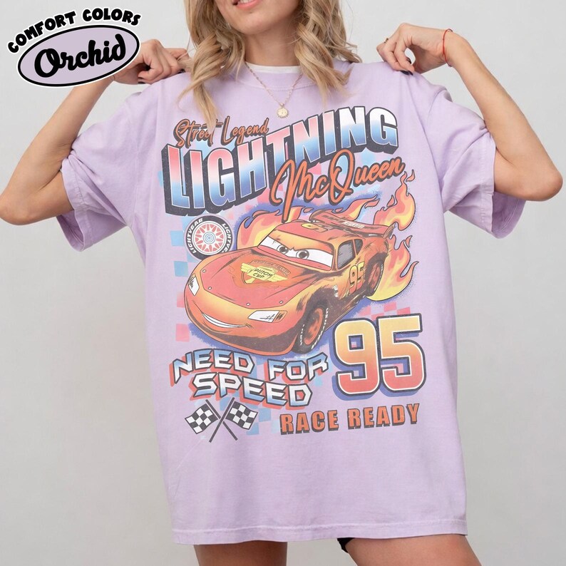 Puede incluir: Una camiseta de color lila claro con un dise&ntilde;o de Lightning McQueen de la pel&iacute;cula Cars. El dise&ntilde;o incluye el texto "Street Legend LIGHTNING McQueen", "Need for Speed" y "Race Ready", junto con el n&uacute;mero "95" y banderas a cuadros. La camiseta est&aacute; etiquetada como "Comfort Colors Orchid".