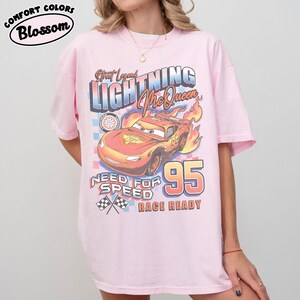 Puede incluir: Una camiseta rosa claro con un dise&ntilde;o de Rayo McQueen de la pel&iacute;cula Cars. El dise&ntilde;o incluye el texto "Street Legend Lightning McQueen", "Need for Speed" y "Race Ready", junto con el n&uacute;mero "95". La camiseta tiene mangas cortas y cuello redondo.
