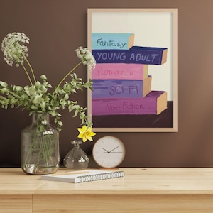 May include: A framed wall art print featuring a stack of five colourful books with the labels Fantasy, Young Adult, Romance, Sci-Fi, and Non-Fiction written on their spines. The books are rendered in shades of blue, dark blue, pink, purple, and magenta.