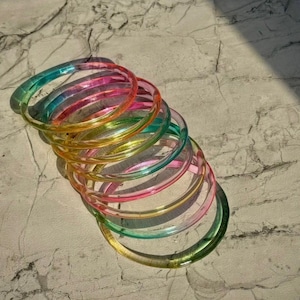 Könnte beinhalten: Ein Stapel bunter, transparenter Armreifen. Die Armreifen sind in den Farbtönen Grün, Gelb, Rosa und Blau gehalten. Die Armbänder liegen auf einer hellgrauen, strukturierten Oberfläche.
