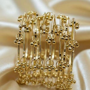 Pode incluir: Um conjunto de pulseiras douradas com um design delicado. Cada pulseira apresenta pequenos grupos pendurados de contas redondas. As pulseiras são dispostas sobre uma superfície de cetim, criando uma exibição luxuosa e elegante.