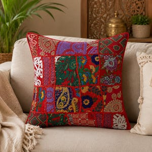 May include: A square patchwork cushion with a red border. The cushion's centre is made up of various fabric squares in red, purple, green, and gold. The cushion is placed on a beige sofa, adding a decorative touch.