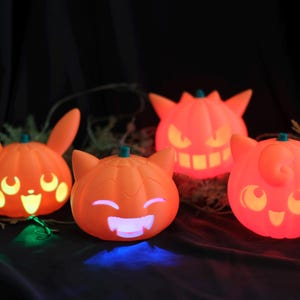 May include: Four orange Halloween-themed light-up decorations. The decorations are shaped like pumpkins with various cartoon character faces. The lights glow in orange, yellow, and white. The background is dark.