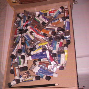 May include: A wooden artist's box filled with numerous tubes of acrylic paint in various colors. The box is open, revealing the paint tubes and a black pencil. The box has a leather handle and metal clasps.