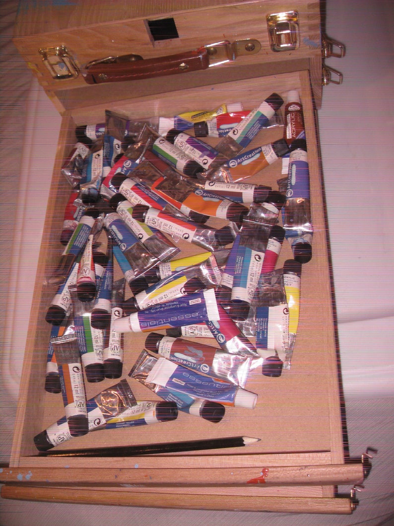 May include: A wooden artist's box filled with numerous tubes of acrylic paint in various colors. The box is open, revealing the paint tubes and a black pencil. The box has a leather handle and metal clasps.